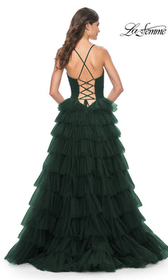 A woman wearing the La Femme Lace-Up Long Tiered Prom Dress 32086 in dark green stands facing away, highlighting the full length, ruffled tiers, and lace-up open back design of the gown.