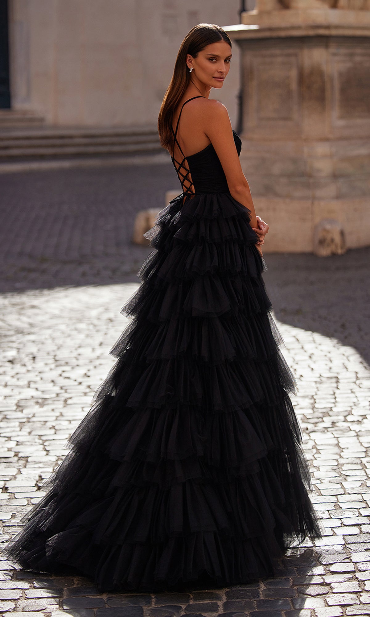 A woman wearing the La Femme Lace-Up Long Tiered Prom Dress 32086 stands on a cobblestone street, gazing over her shoulder with her hair down.