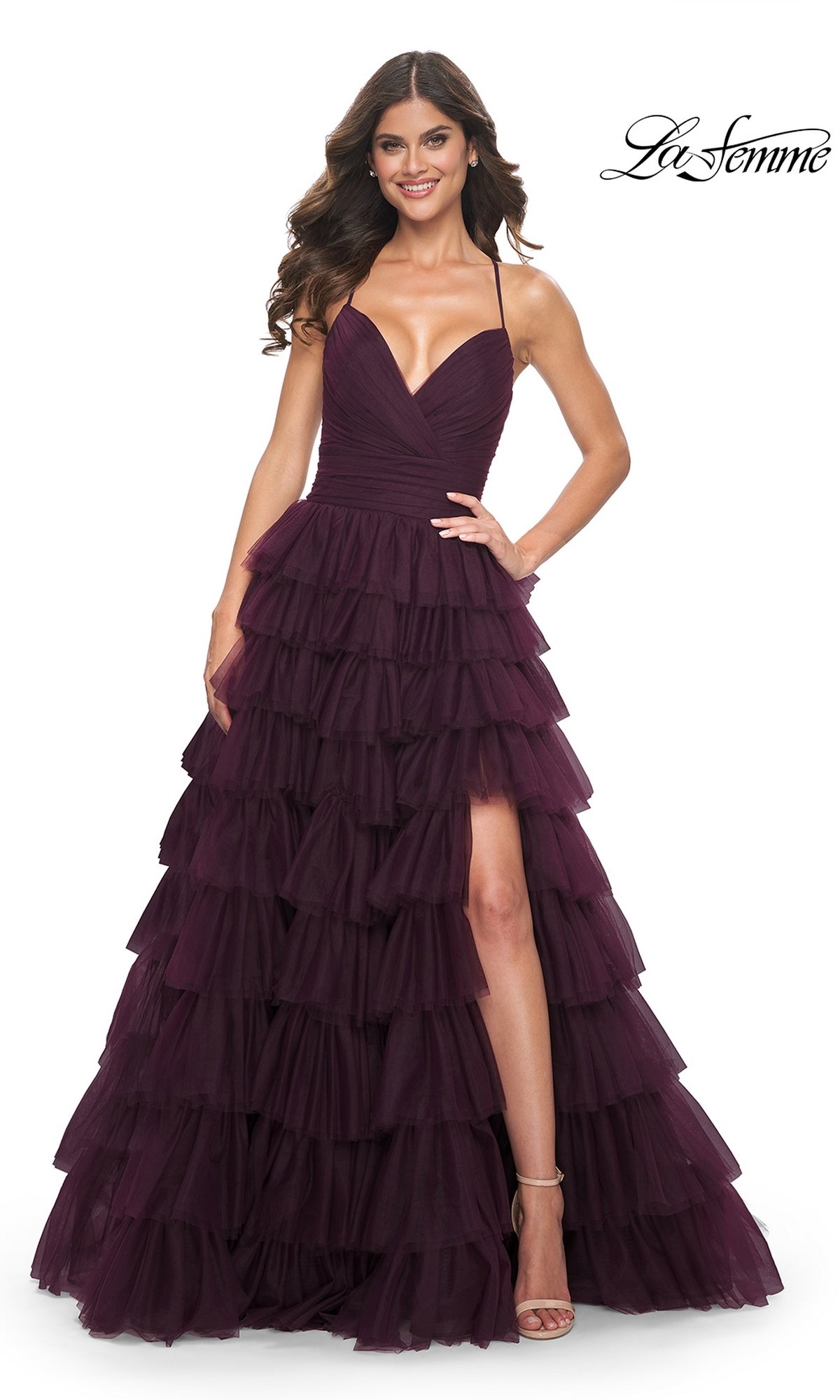 A woman poses and smiles in the La Femme Lace-Up Long Tiered Prom Dress 32086, featuring a dark purple hue, deep V-neckline, thin straps, high leg slit, and elegant tulle layers against a white background.