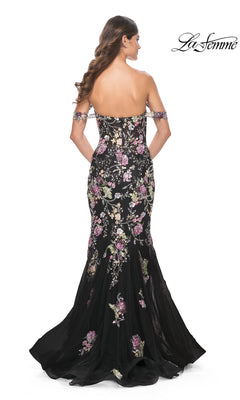 Woman in the La Femme Long Floral Mermaid Prom Dress 32087, a black floor-length off-the-shoulder gown with floral embroidery, shown from the back with loose wavy hair. "La Femme" logo appears in the top right corner.