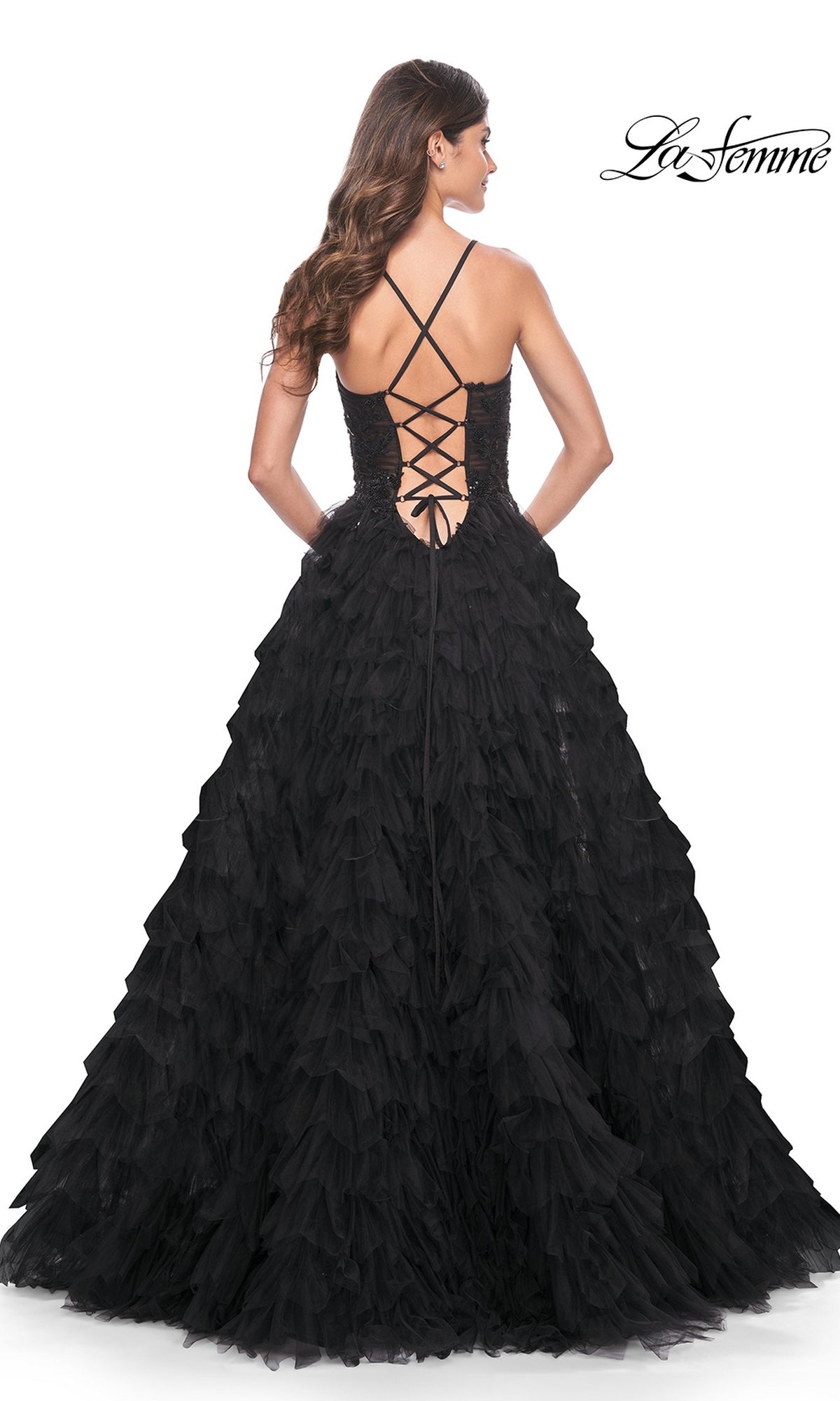 A woman stands with her back to the camera in the La Femme Long Black Ruffled Prom Ball Gown 32108, featuring a lace-up back, crisscross straps, and a dramatic floor-length A-line silhouette.