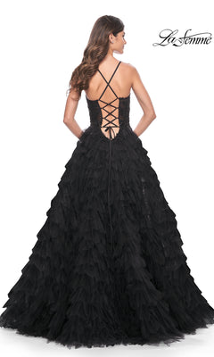 A woman stands with her back to the camera in the La Femme Long Black Ruffled Prom Ball Gown 32108, featuring a lace-up back, crisscross straps, and a dramatic floor-length A-line silhouette.