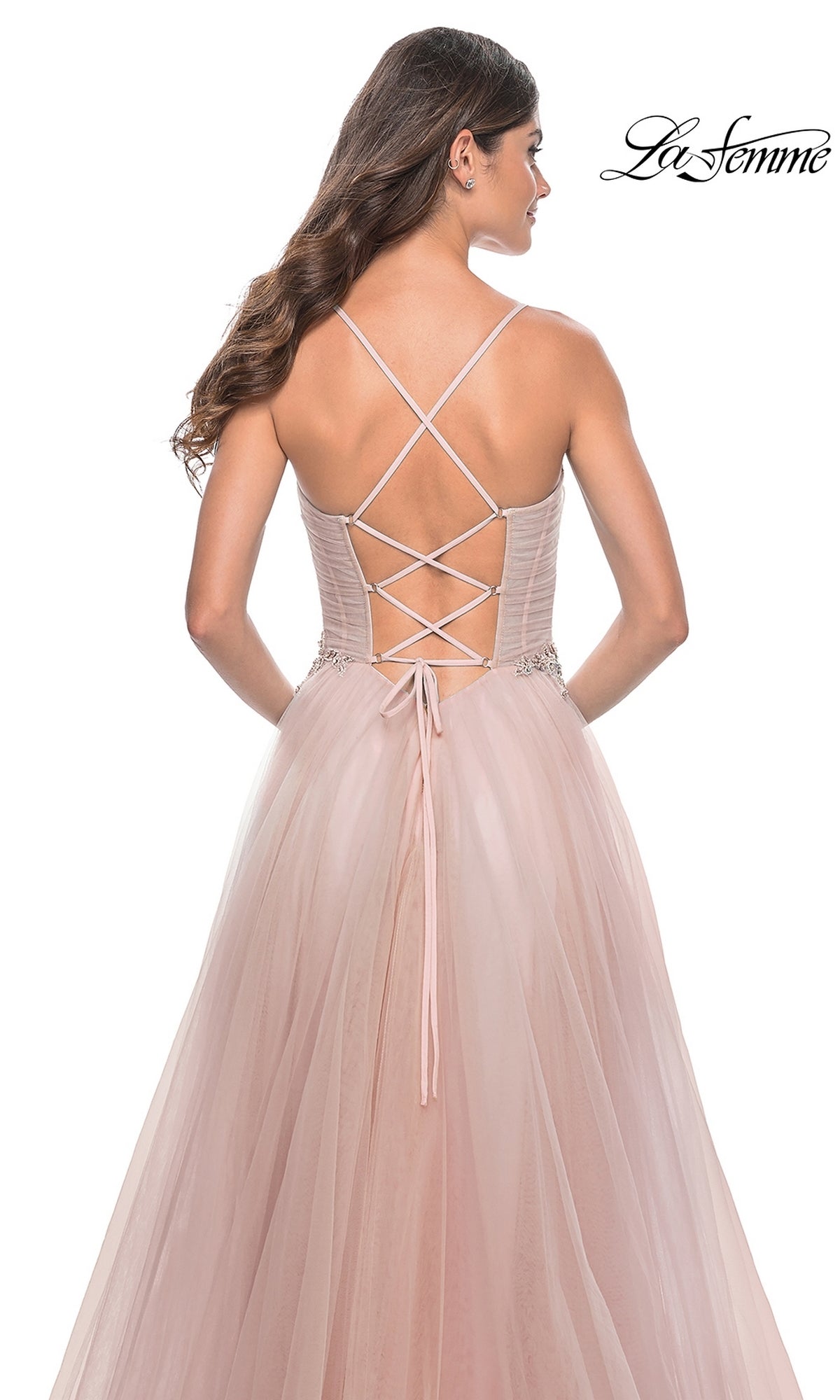 Woman in a blush pink La Femme Sheer-Bodice Long A-Line Prom Dress 32117 with strappy open-back, thin crisscross straps, tulle skirt, and beaded waist detail, standing with her back to the camera.