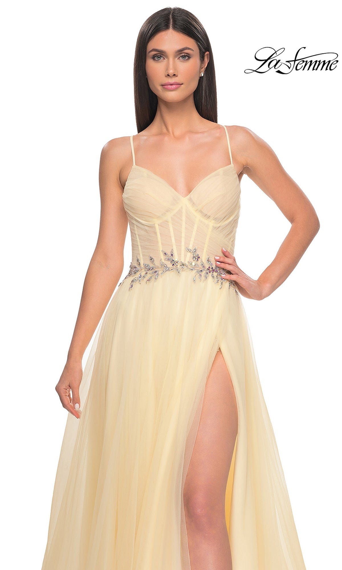 A woman in a light yellow La Femme Sheer-Bodice Long A-Line Prom Dress 32117 with thin straps, floral waist embellishments, and a high slit stands against a white background. The 