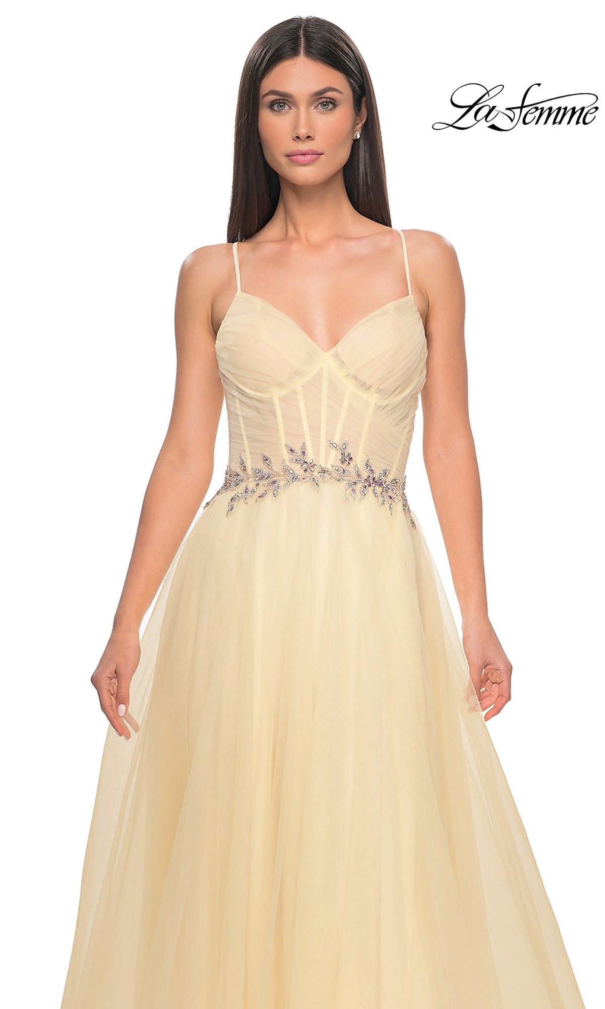 A woman wears the La Femme Sheer-Bodice Long A-Line Prom Dress 32117—a light yellow, sleeveless gown with thin straps, floral embroidery at the waist, and a flowing tulle skirt. 