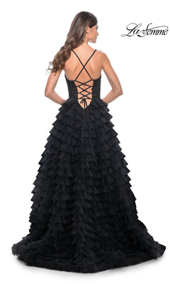 Woman wearing the La Femme Long Ruffled Prom Ball Gown 32128 in black, featuring a tiered skirt, lace-up back, and thin crisscross straps, standing and facing away from the camera.