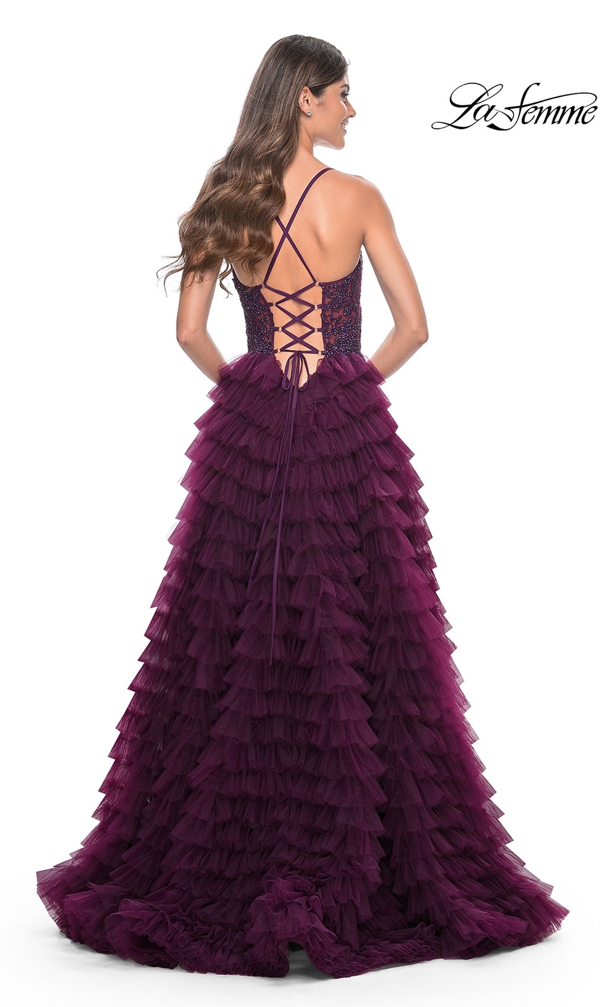 A woman stands with her back to the camera in the La Femme Long Ruffled Prom Ball Gown 32128, a deep purple formal dress with floor-length tiered ruffles and a lace-up open back. The 