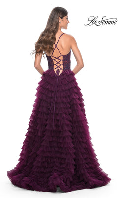 A woman stands with her back to the camera in the La Femme Long Ruffled Prom Ball Gown 32128, a deep purple formal dress with floor-length tiered ruffles and a lace-up open back. The "La Femme" logo is visible in the top right corner.