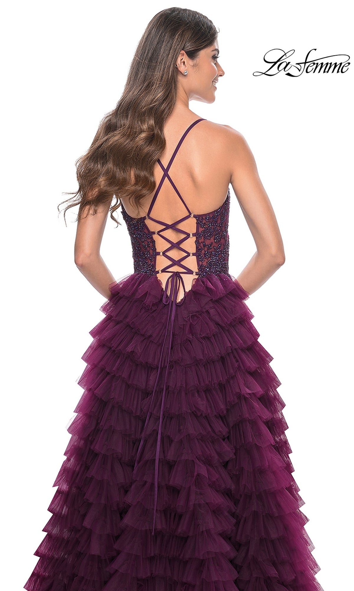 A woman stands with her back to the camera in the La Femme Long Ruffled Prom Ball Gown 32128, an elegant deep purple A-line dress featuring a lace-up back and a layered ruffled skirt.