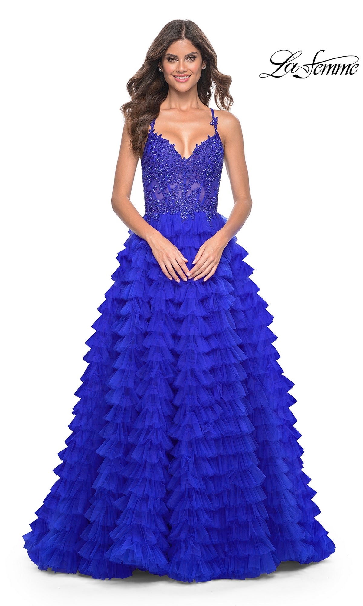 A woman wears the La Femme Long Ruffled Prom Ball Gown 32128 featuring a bright blue color, fitted lace bodice, spaghetti straps, and ruffled skirt, standing against a white background.