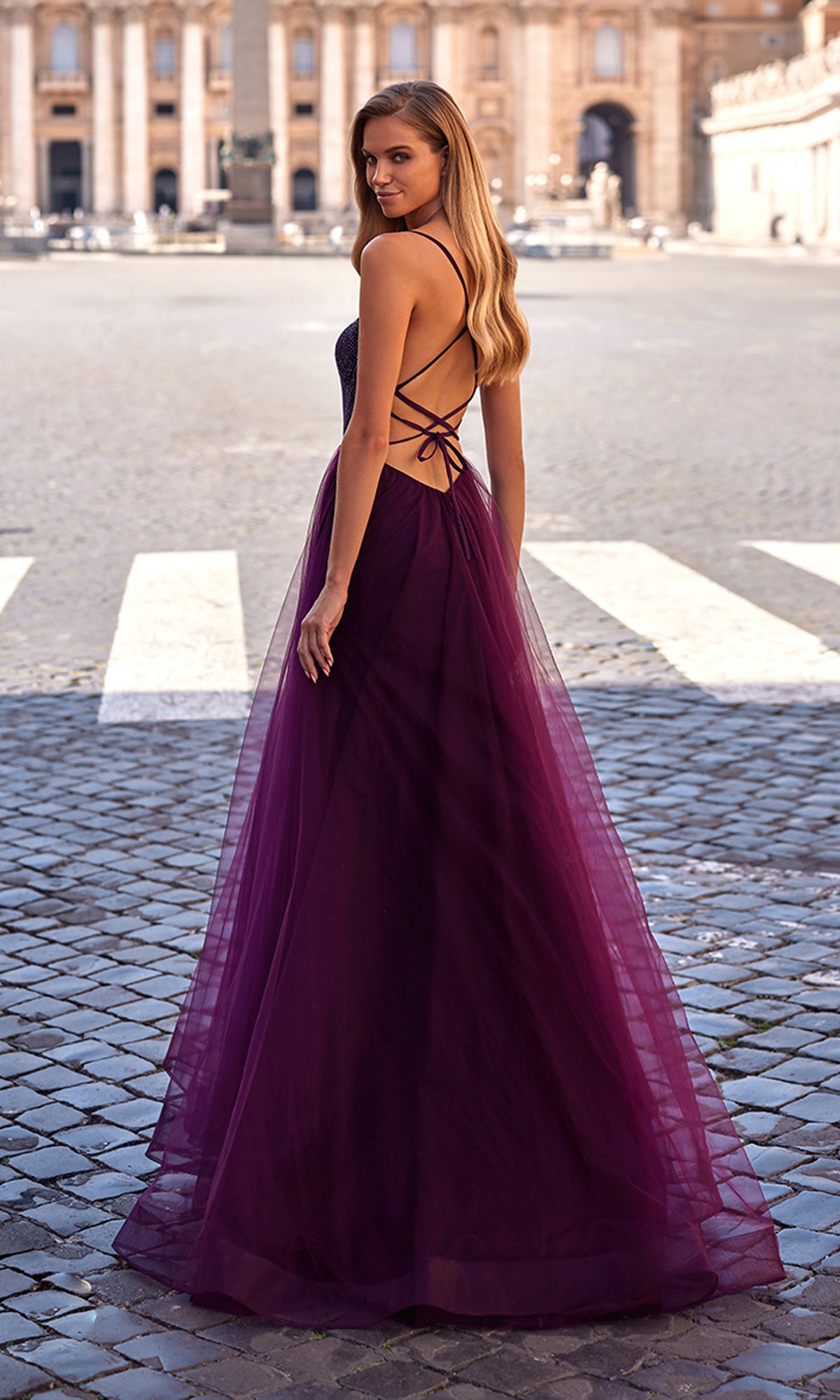 A woman in the La Femme Embellished Long Tulle Prom Dress 32135, featuring a backless design with a beaded bodice and flowing deep purple skirt, stands outdoors on cobblestone pavement, glancing over her shoulder.
