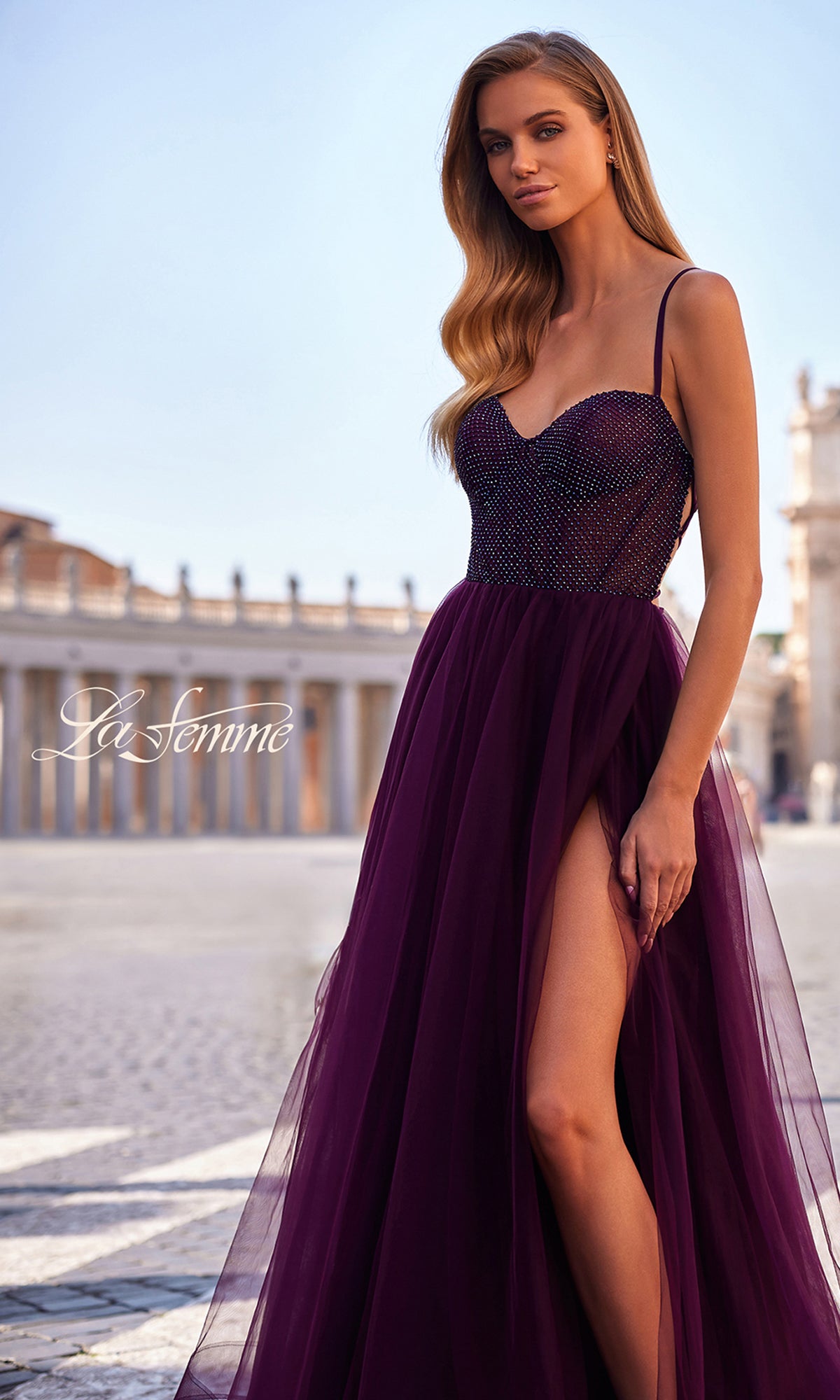 A woman in the La Femme Embellished Long Tulle Prom Dress 32135 stands outdoors on a paved area, classical architecture in the background.