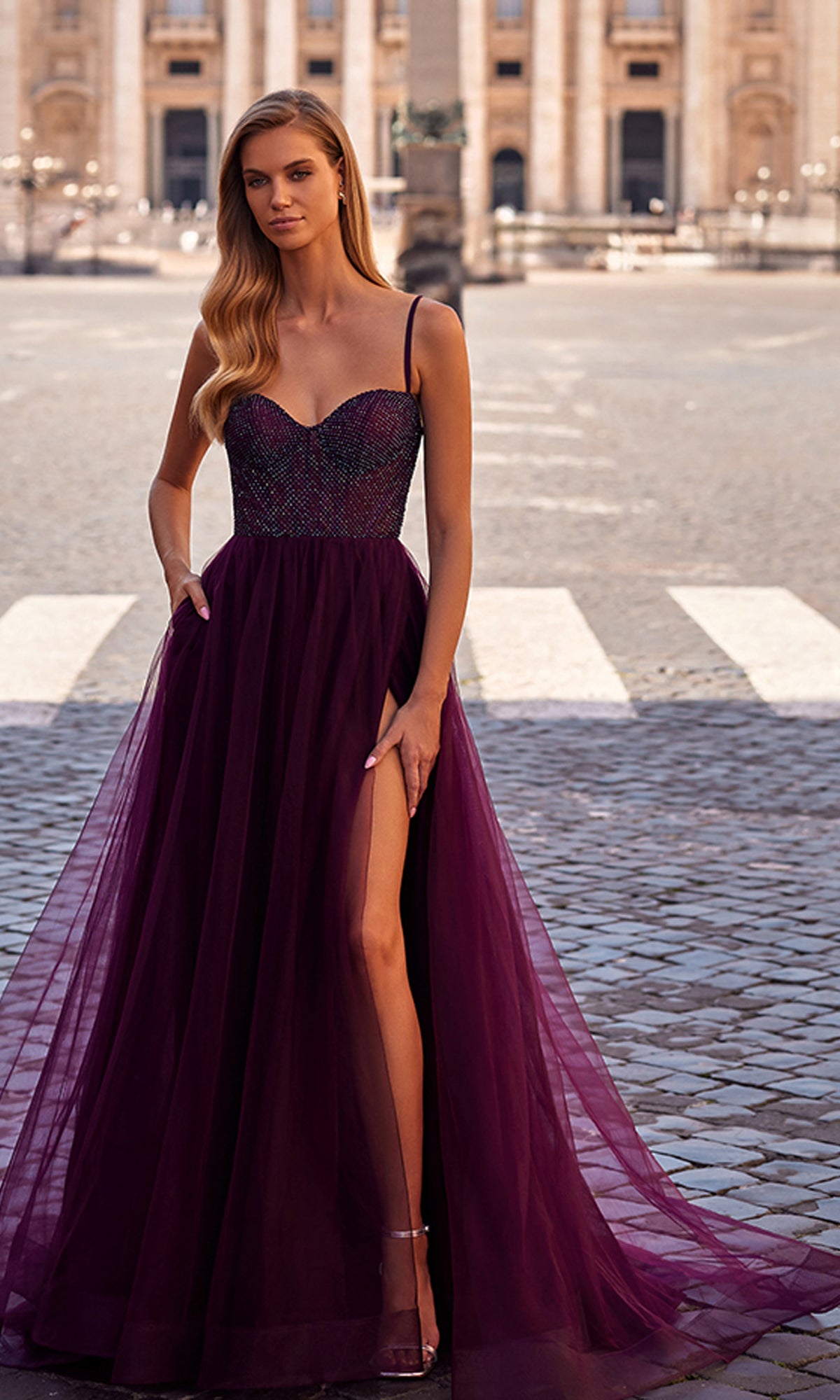 Woman in a La Femme Embellished Long Tulle Prom Dress 32135 in Dark Berry, featuring a sleeveless beaded bodice and sheer skirt with thigh-high slit, standing on a cobblestone street against a blurred city background.