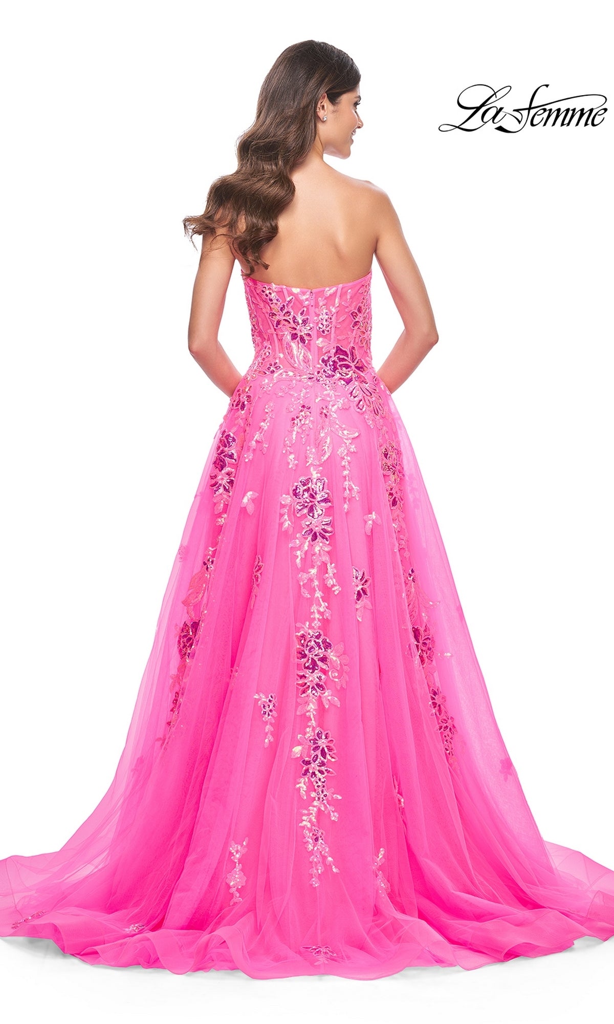 A woman with long brown hair models the La Femme Neon Pink Long Prom Ball Gown 32137, a strapless dress with sequin floral embroidery, standing with her back to the camera. The La Femme logo appears in the upper right corner.