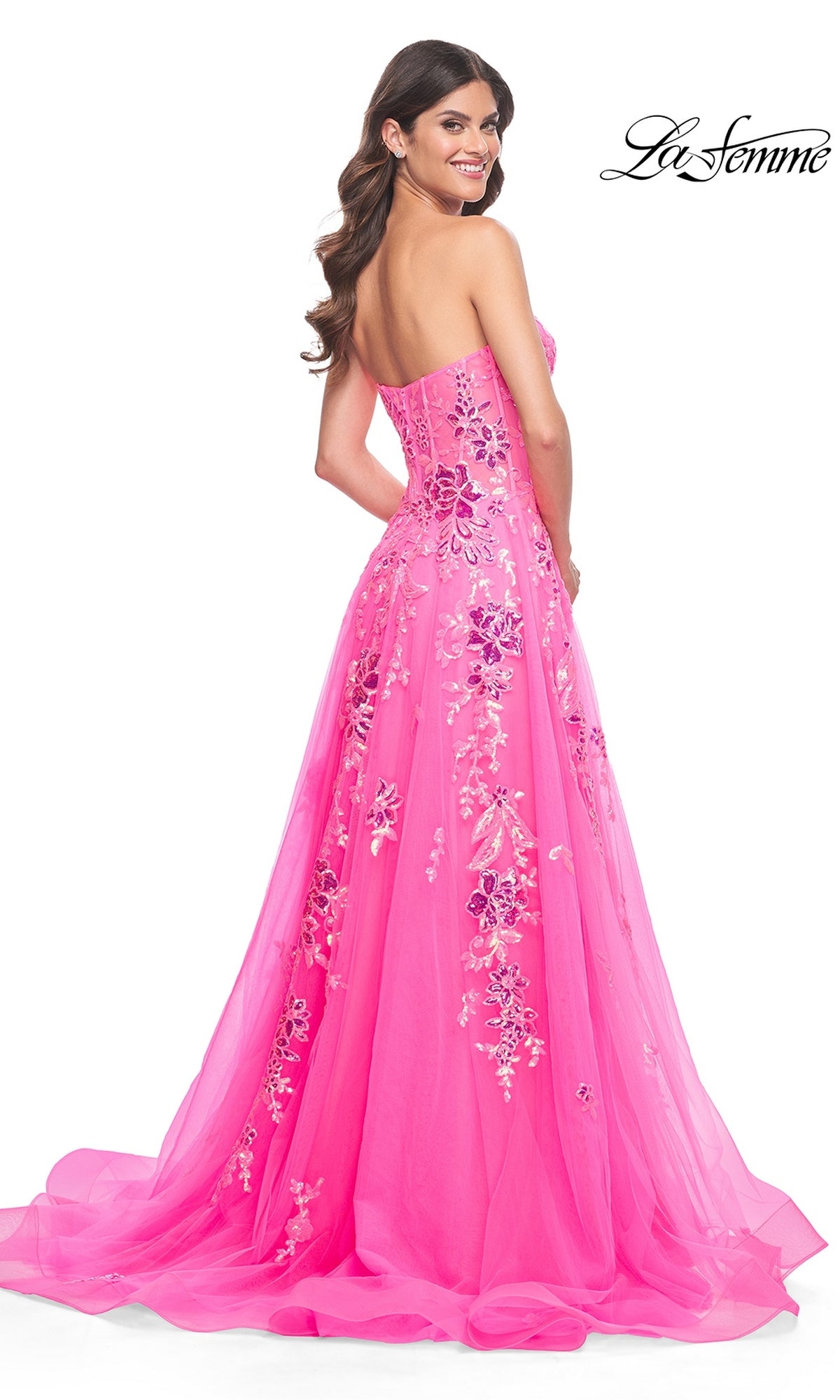 Model wears the La Femme Neon Pink Long Prom Ball Gown 32137, a strapless dress with floral embroidery, posing and smiling over her shoulder against a white background.