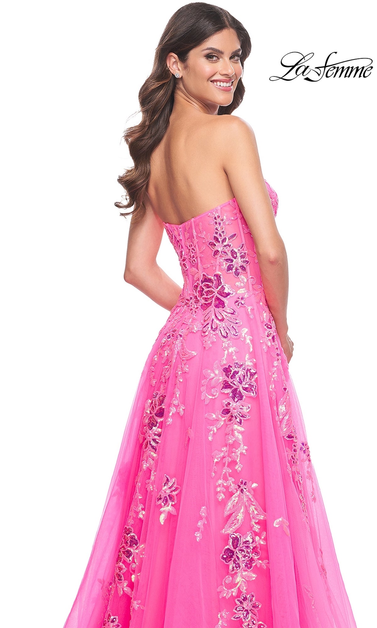 A woman in the La Femme Neon Pink Long Prom Ball Gown 32137, featuring a strapless design and sequin floral embroidery, smiles over her shoulder. The 