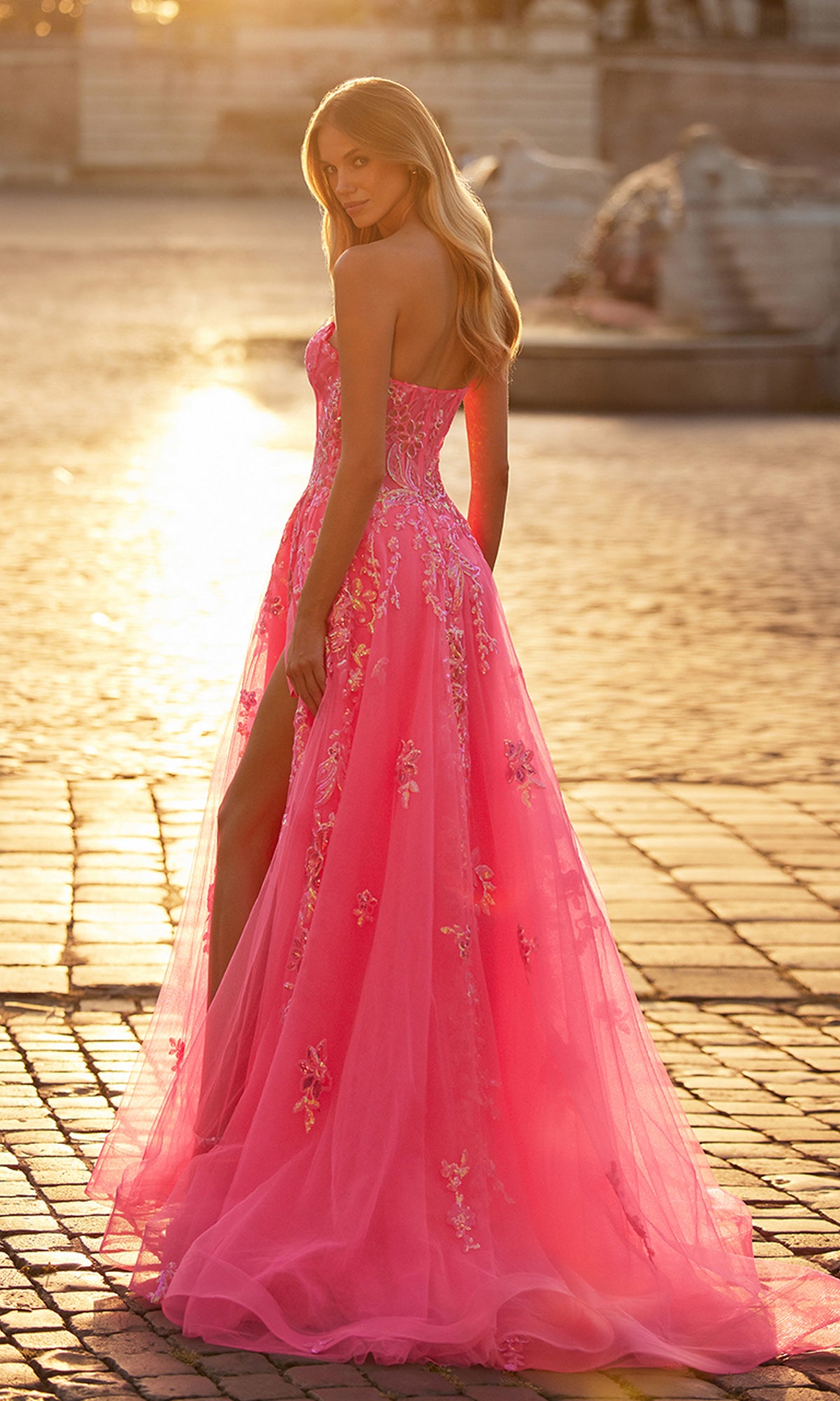 A woman wearing the La Femme Neon Pink Long Prom Ball Gown 32137 with floral embroidery and a high slit stands on a sunlit cobblestone street, glancing over her shoulder.