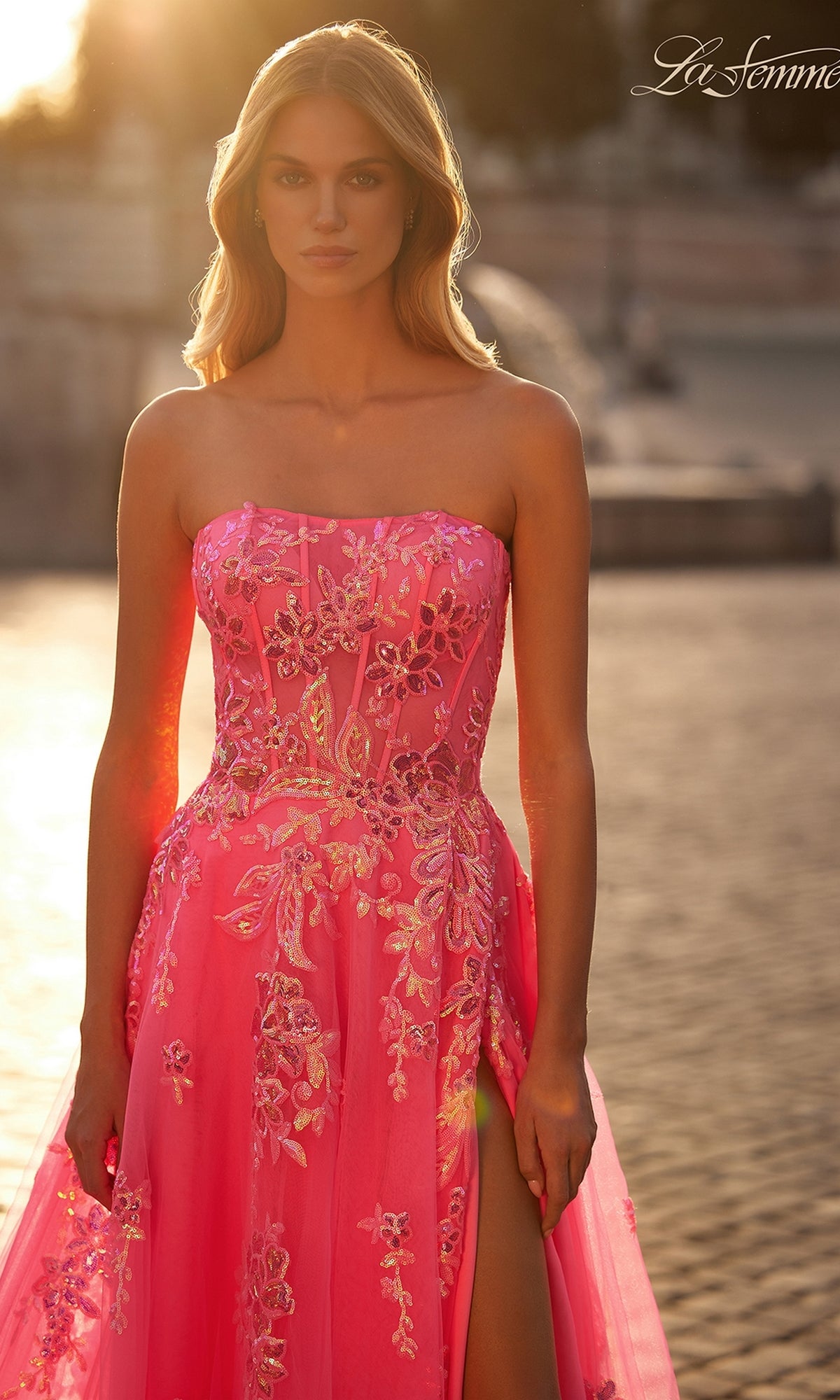 A woman stands outdoors in soft sunlight, wearing the La Femme Neon Pink Long Prom Ball Gown 32137, a strapless formal dress featuring floral embroidery, sequins, and a high leg slit.
