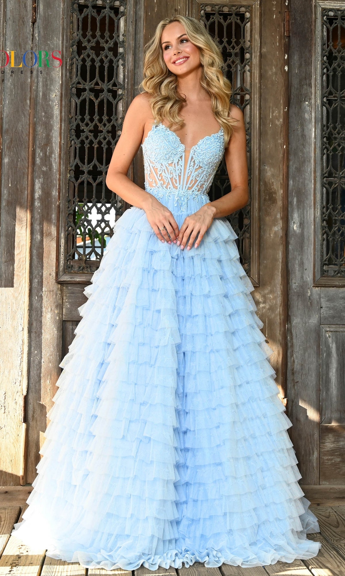 A woman in the Beaded-Corset Long Ruffled Prom Ball Gown 3219 with a sheer lace bodice poses, smiling slightly and looking left, in front of an intricately carved wooden door.