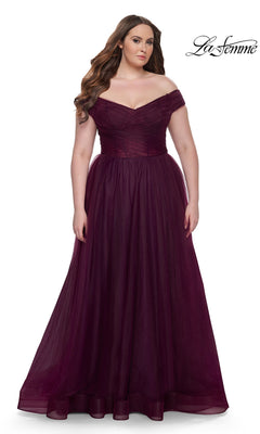 A woman in the La Femme Plus-Size Long A-Line Prom Dress 32204 stands against a white background. The formal gown features an off-the-shoulder fitted bodice and flowing skirt. The "La Femme" logo appears in the top right corner.