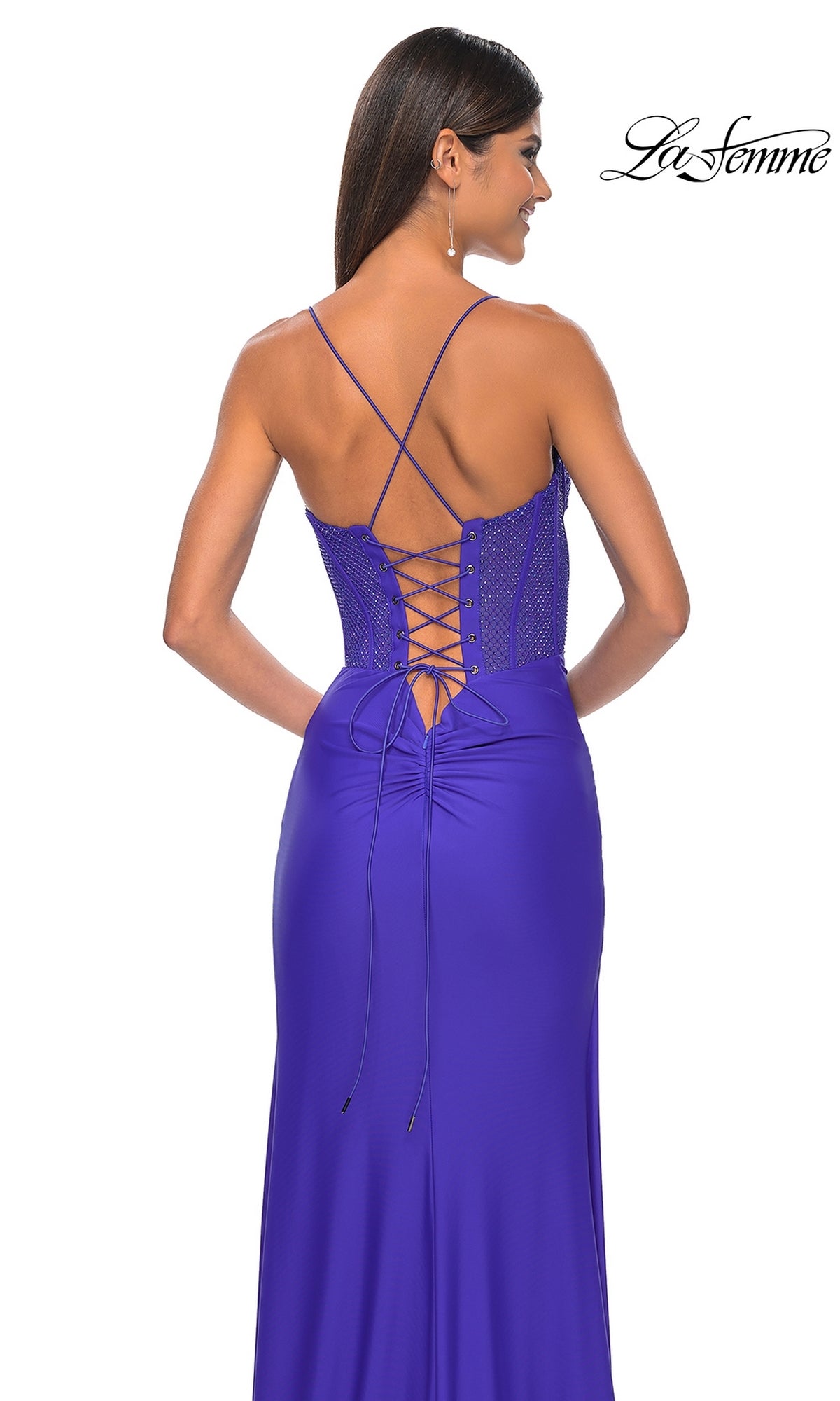A woman wearing the La Femme Lace-Up Long Fishnet Prom Dress 32230 in royal blue, featuring a sleeveless design, thin straps, sheer corset bodice, and lace-up back, stands facing away from the camera.
