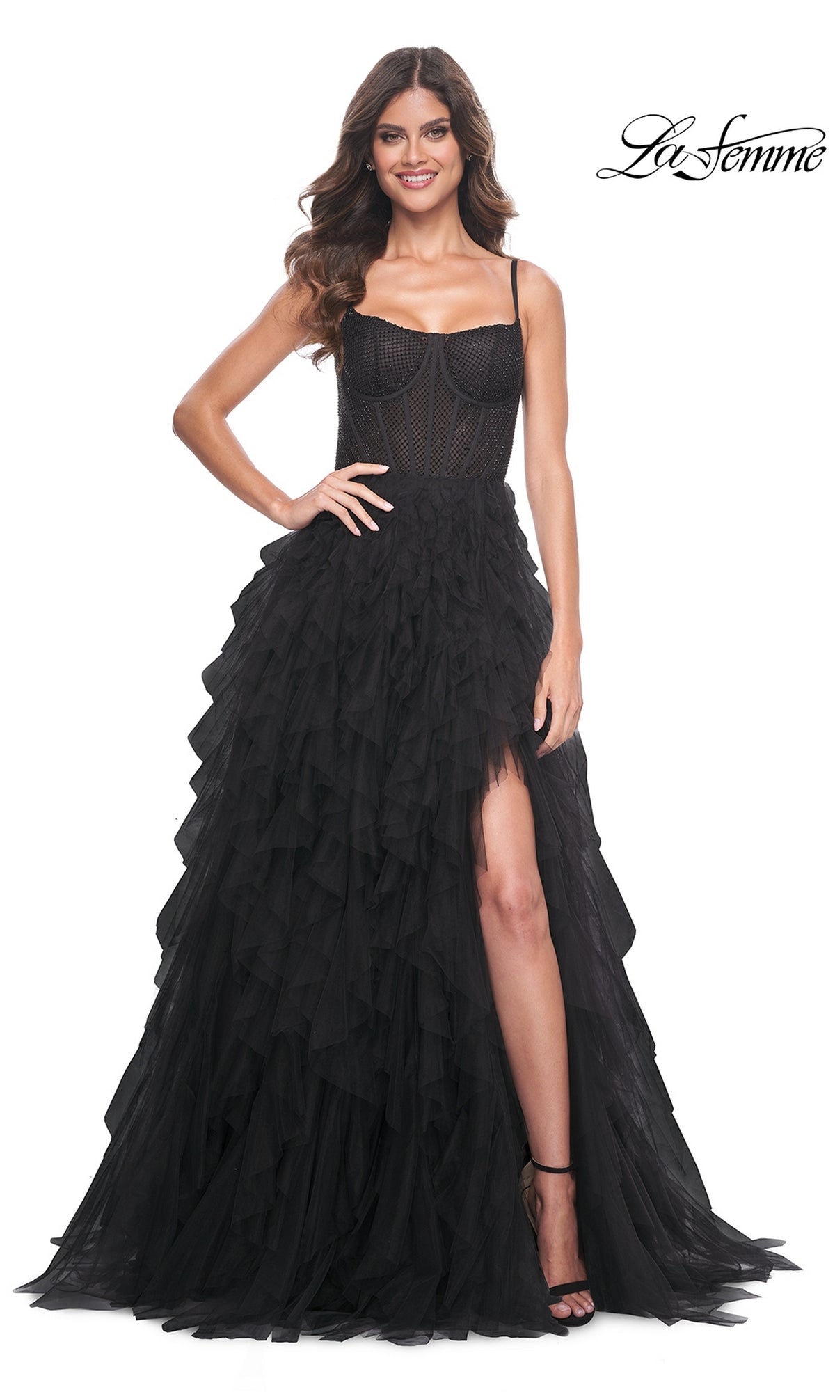 A woman wearing the La Femme Sheer-Waist Long Ruffled Prom Dress 32233 in black, featuring a corset-style bodice and thigh-high slit, stands smiling against a white background with the 