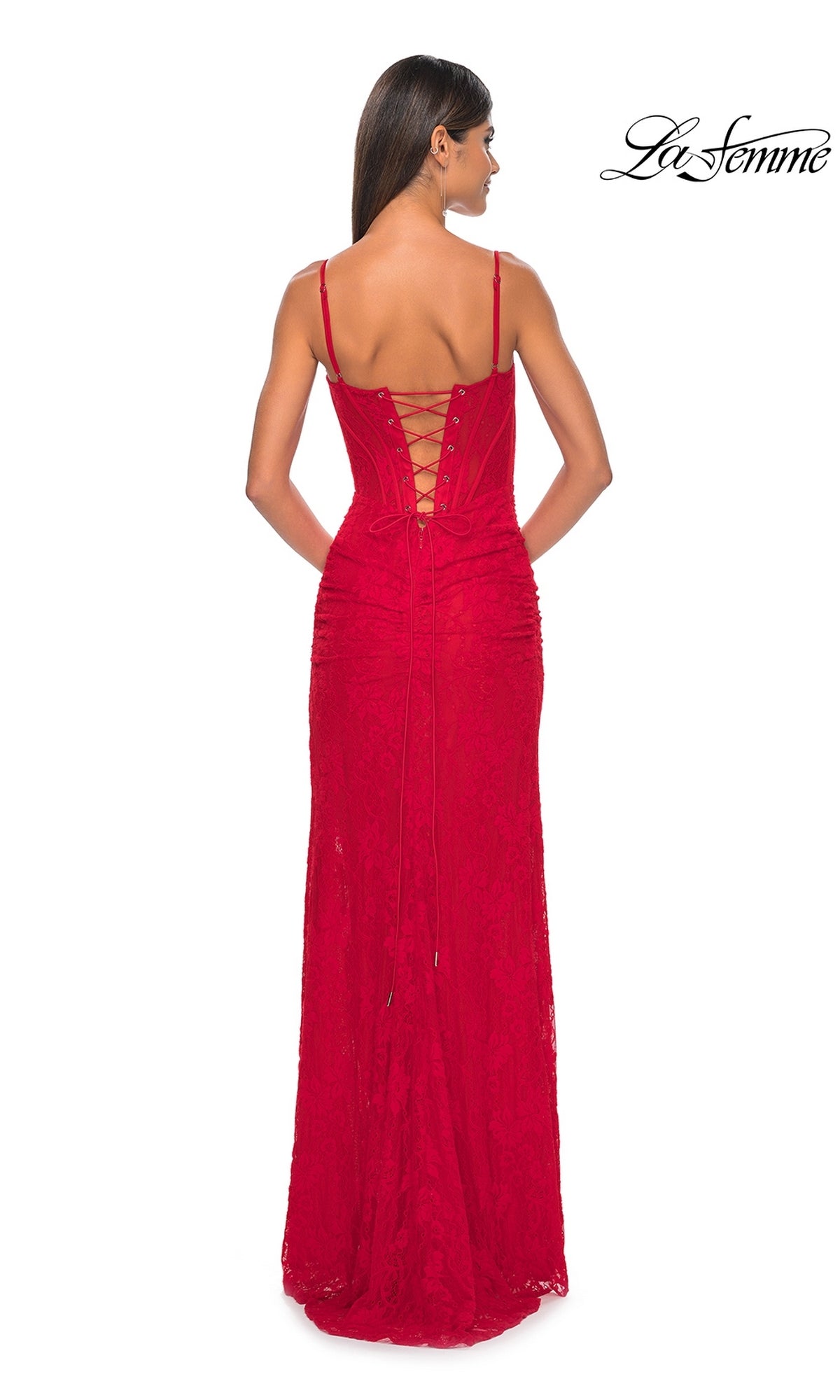 A woman models the La Femme Lace-Up Back Long Lace Prom Dress 32237, a red lace designer gown with thin straps, a corset-style bodice, and lace-up back detail, standing facing away from the camera.