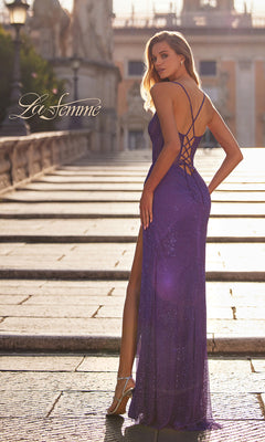 A woman in the La Femme Beaded-Fishnet Long Prom Dress 32247, featuring a purple backless design with sequins and a high slit, stands on a sunlit stone walkway, looking over her shoulder. "La Femme" appears on the left side.