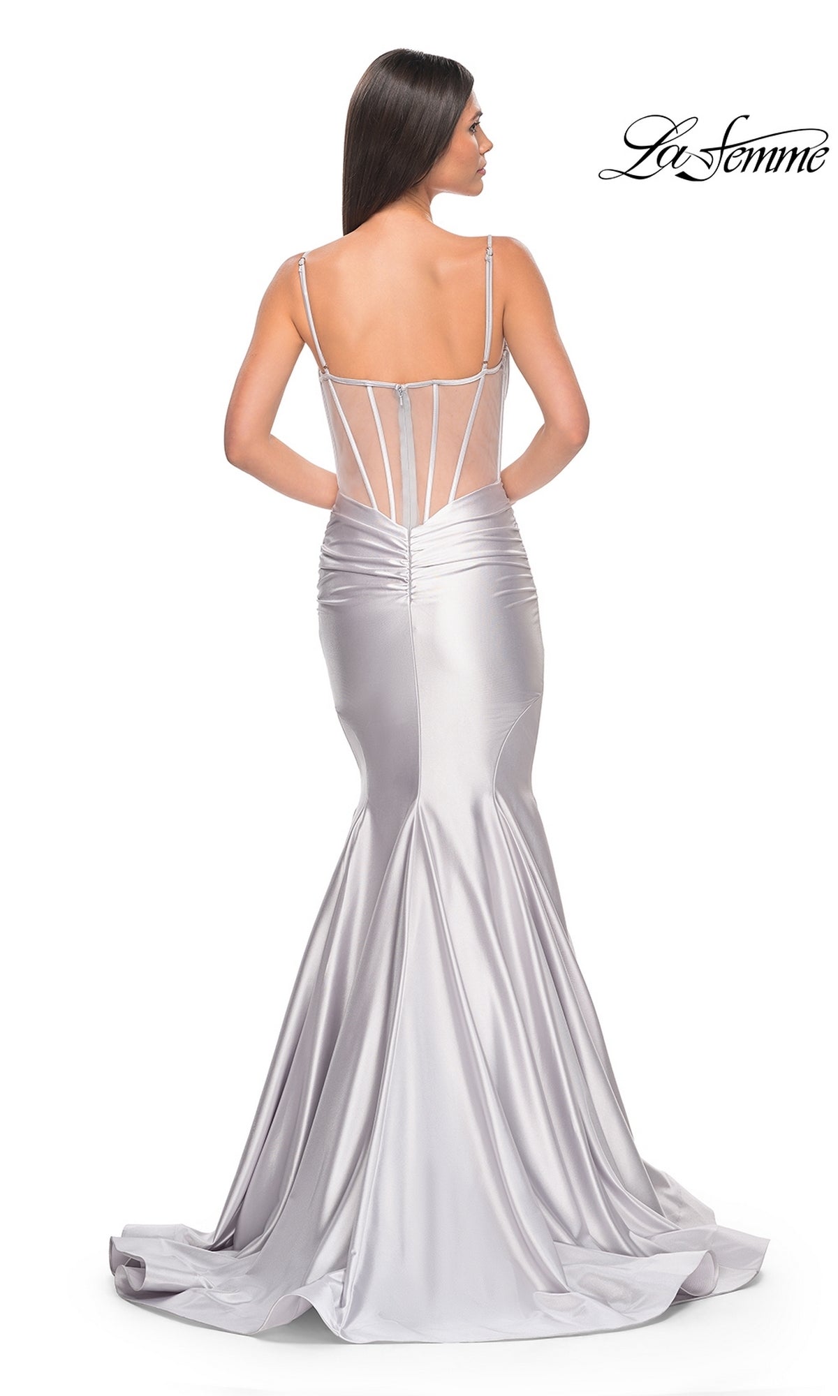 A woman in the La Femme Long Corset Mermaid Prom Dress 32269, featuring silver satin fabric, thin straps, sheer panels, and a long train, stands with her back to the camera.