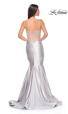 A woman in the La Femme Long Corset Mermaid Prom Dress 32269, featuring silver satin fabric, thin straps, sheer panels, and a long train, stands with her back to the camera.
