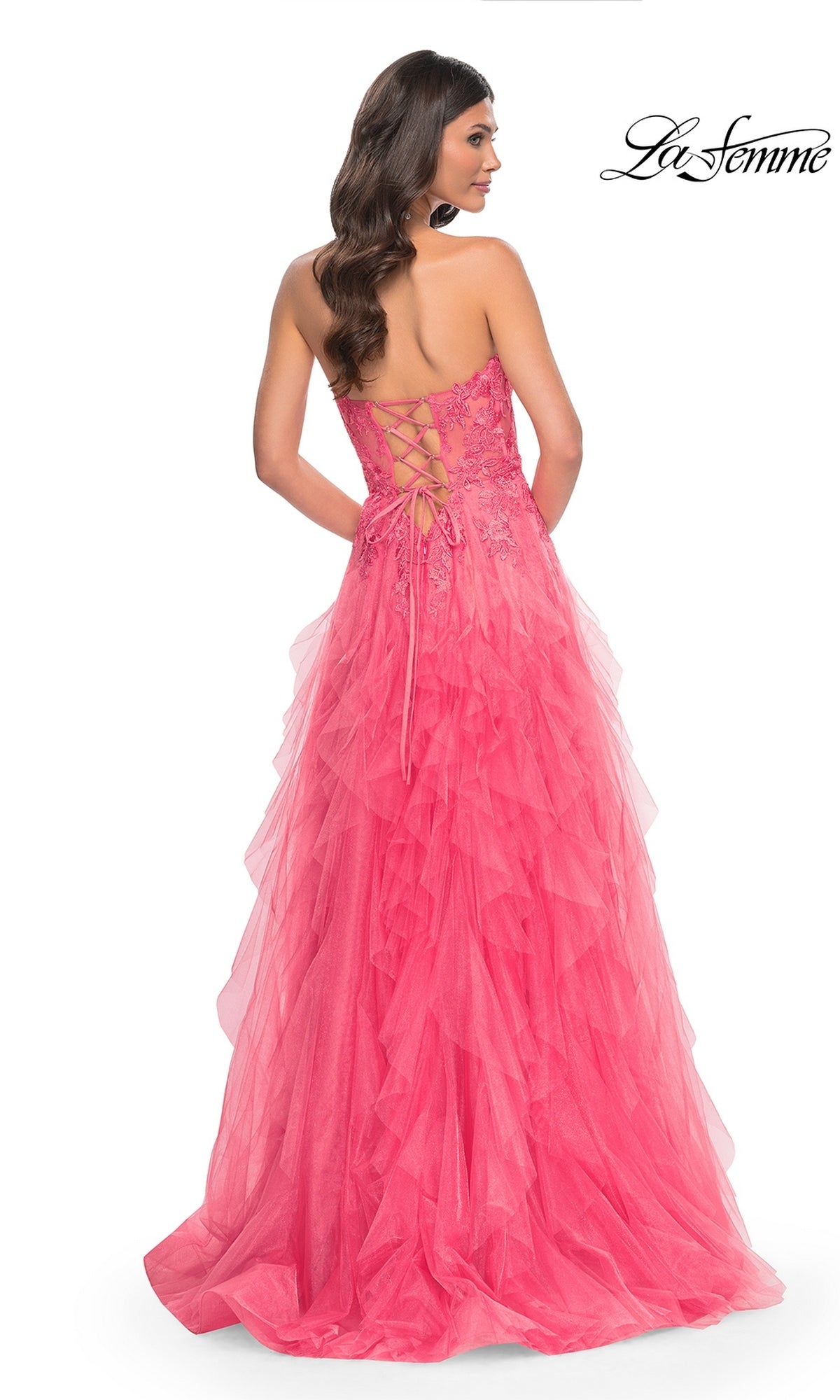 A woman models the La Femme Strapless Long Coral Prom Dress 32286, featuring a beaded-lace bodice, lace-up back, and flowy floor-length tulle skirt, as she stands facing away from the camera.