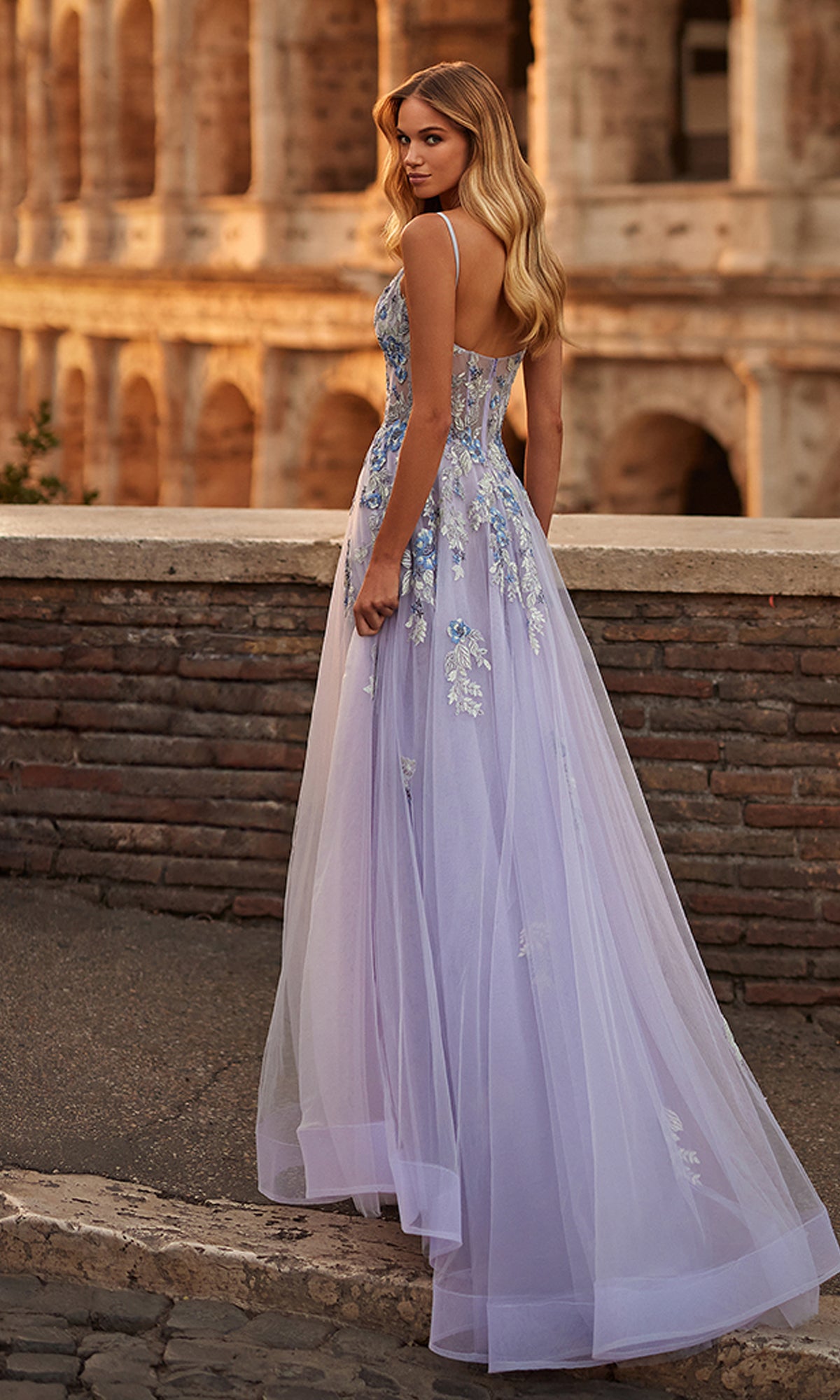 A woman wears the La Femme Long Lavender A-Line Prom Dress 32288 with floral embroidery, posing outdoors by a stone railing in front of an ancient Roman amphitheater.