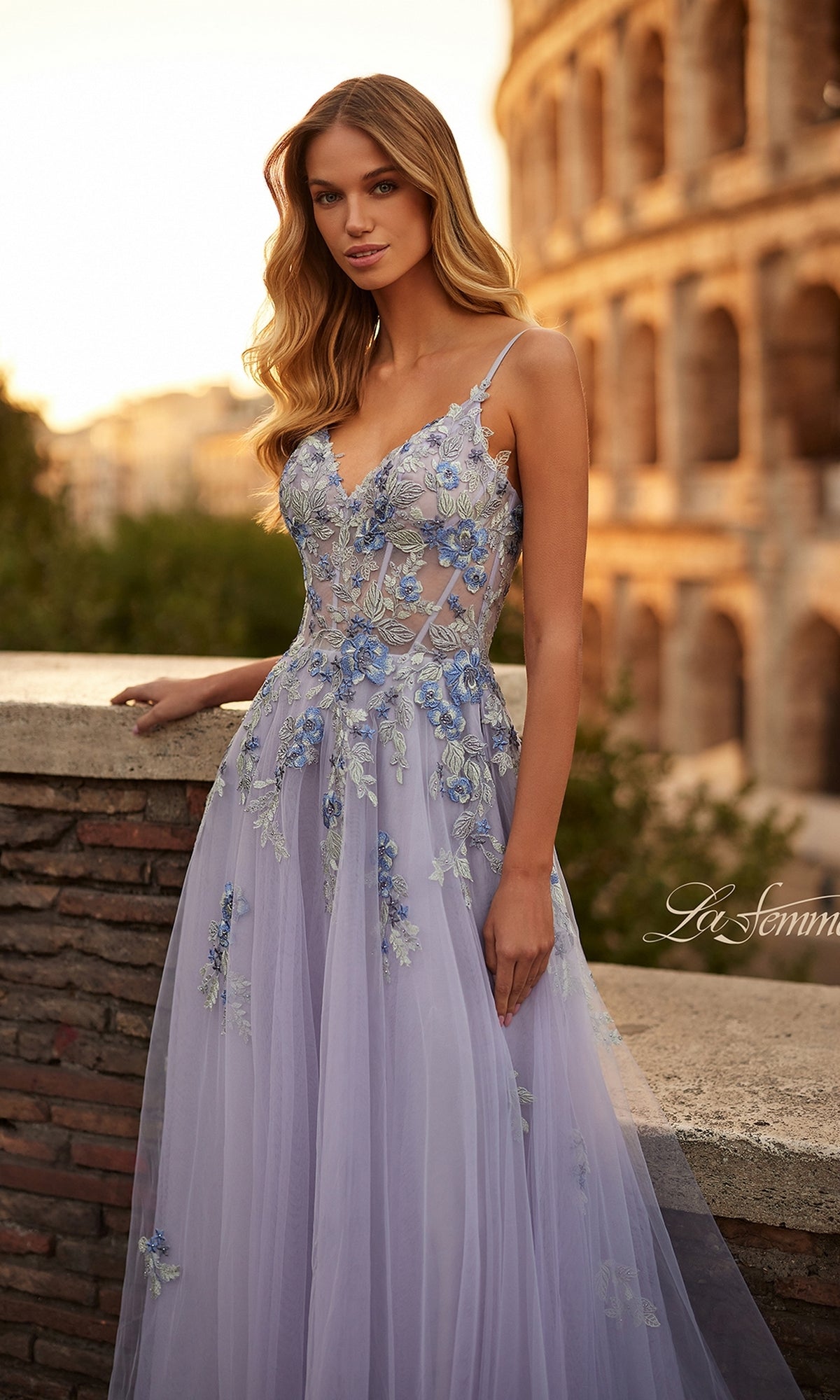 A woman in the La Femme Long Lavender A-Line Prom Dress 32288 with floral embroidery stands by a stone wall as the Colosseum glows at sunset behind her.