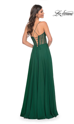 A woman stands facing away in the La Femme Sheer-Waist Long A-Line Prom Dress 32296, featuring an emerald green floor-length skirt, lace-up corset bodice, and sheer side panels.
