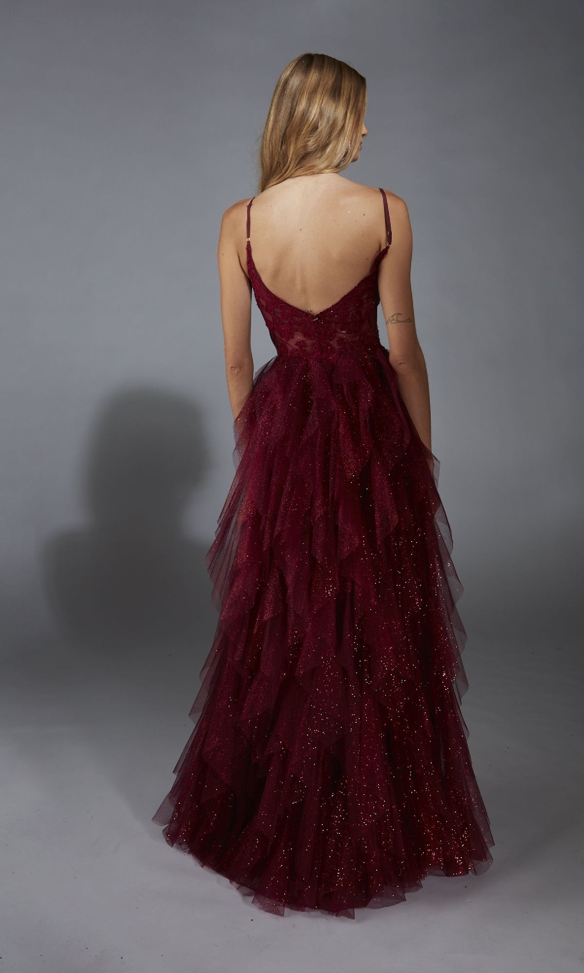 A person with long blonde hair wears the Aerie Couture 322 Glitter Ruffle Prom Ball Gown—deep red, glittery, thin straps, tiered skirt with leg slit—shown from the back against a gray background.