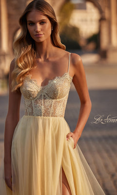 A woman in a sheer yellow La Femme Lace-Bodice Long A-Line Prom Dress 32306 stands outdoors on a cobblestone path, with an arched stone structure in the background.