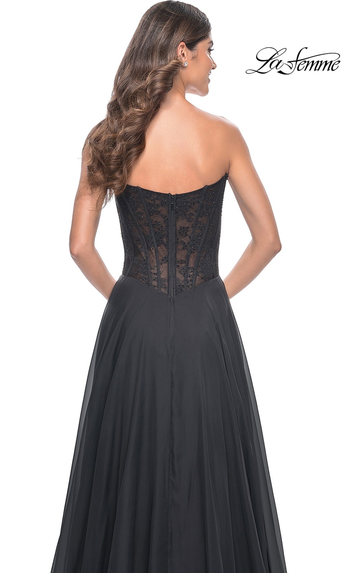 A woman with long, wavy brown hair is seen from the back in the black La Femme Lace-Bodice Long A-Line Prom Dress 32311, featuring a strapless lace corset bodice. The 