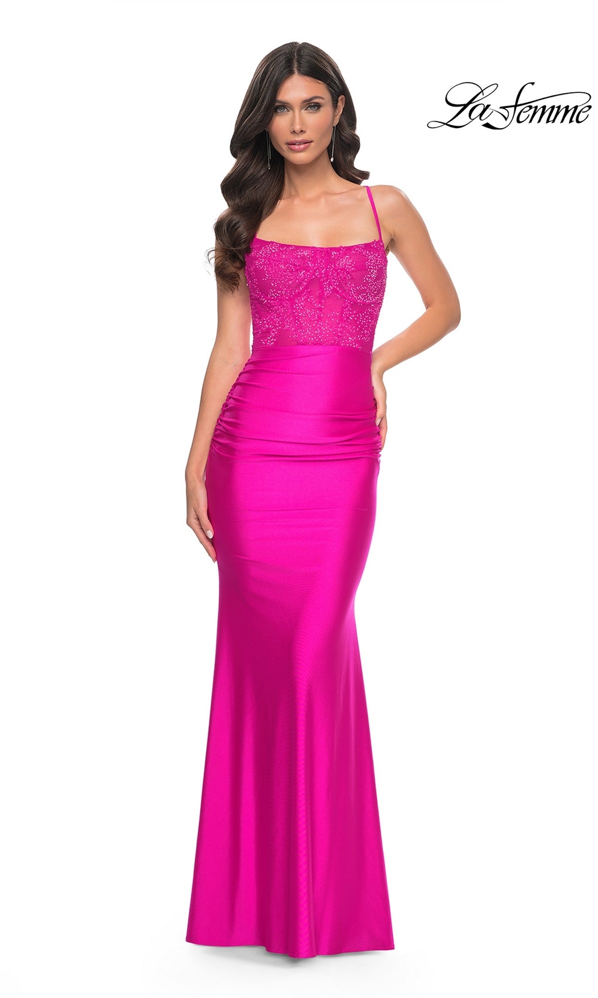 A woman in the La Femme Sheer-Bodice Long Prom Dress 32322, featuring a bright pink hue, thin straps, and lace detailing on the bodice, stands against a white background.
