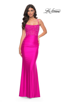 A woman in the La Femme Sheer-Bodice Long Prom Dress 32322, featuring a bright pink hue, thin straps, and lace detailing on the bodice, stands against a white background.