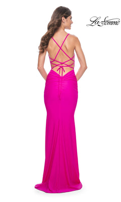 A woman wears the La Femme Backless Bright Long Prom Dress 32325, a floor-length pink gown with a ruched skirt and multiple crisscross straps across the open back.