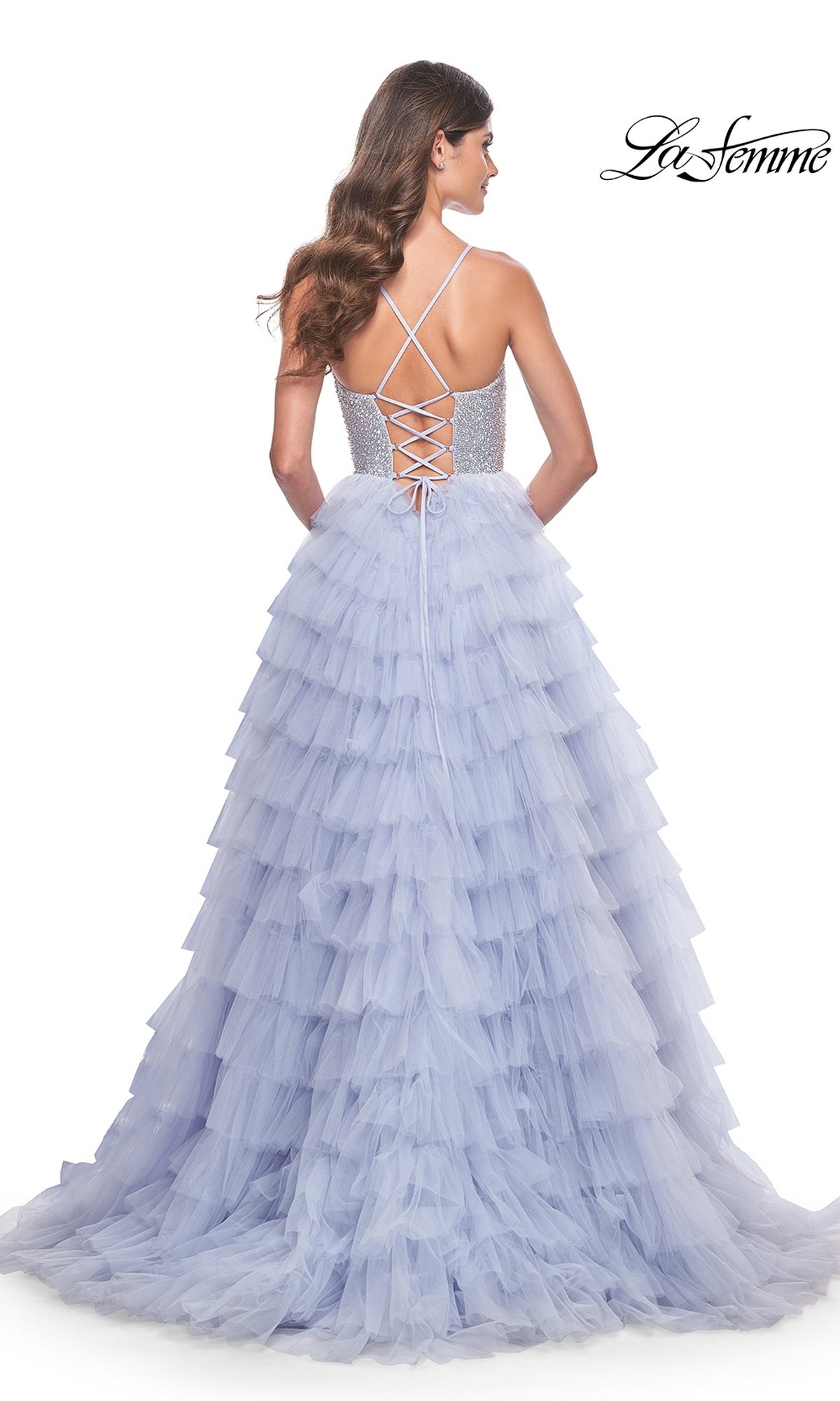 A woman stands facing away in the La Femme Long Tiered A-Line Prom Dress 32335, a lavender gown with a lace-up open back and beaded sweetheart corset detail. The 