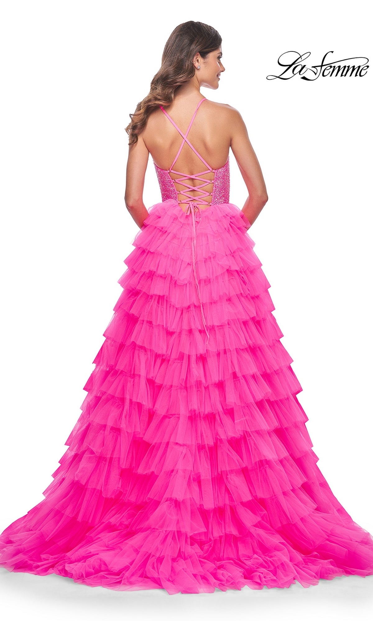 A woman stands facing away in the La Femme Long Tiered A-Line Prom Dress 32335—bright pink with a lace-up back, crisscross straps, beaded sweetheart corset, and a dramatic floor-length tiered skirt.