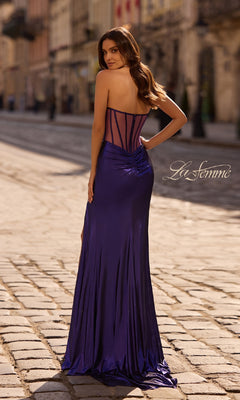 A woman stands on a cobblestone street, facing away, wearing the Strapless Long Metallic Prom Dress La Femme 32375 in deep blue satin with a sheer corset back. The "La Femme" logo is visible on the image.