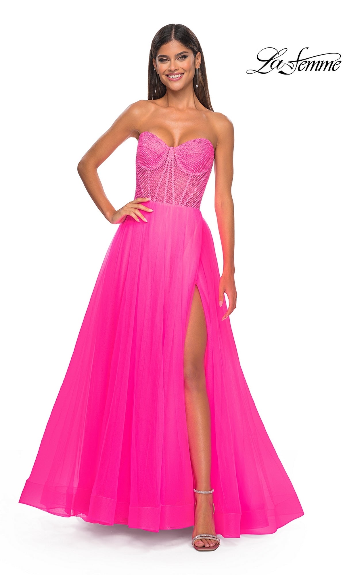 A woman smiles against a white background in the La Femme 32445, a strapless long A-line prom dress in bright pink with a sheer corset bodice and high leg slit.