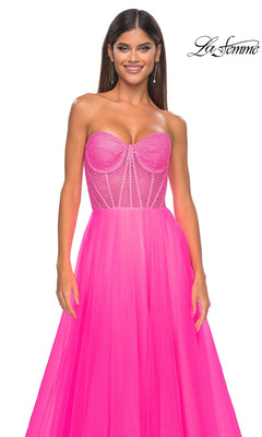 A woman smiles against a white background in the La Femme 32445 strapless long A-line prom dress, featuring a bright pink hue with a fitted corset-style bodice and sheer overlay. The “La Femme” logo appears in the top right corner.