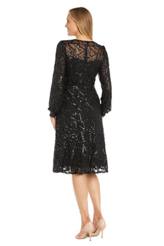 A woman faces away in the Petite Short Black Party Dress: R&M Richards 3250P, a knee-length cocktail dress with long sleeves and black sequins, paired with nude high heels against a plain white background.