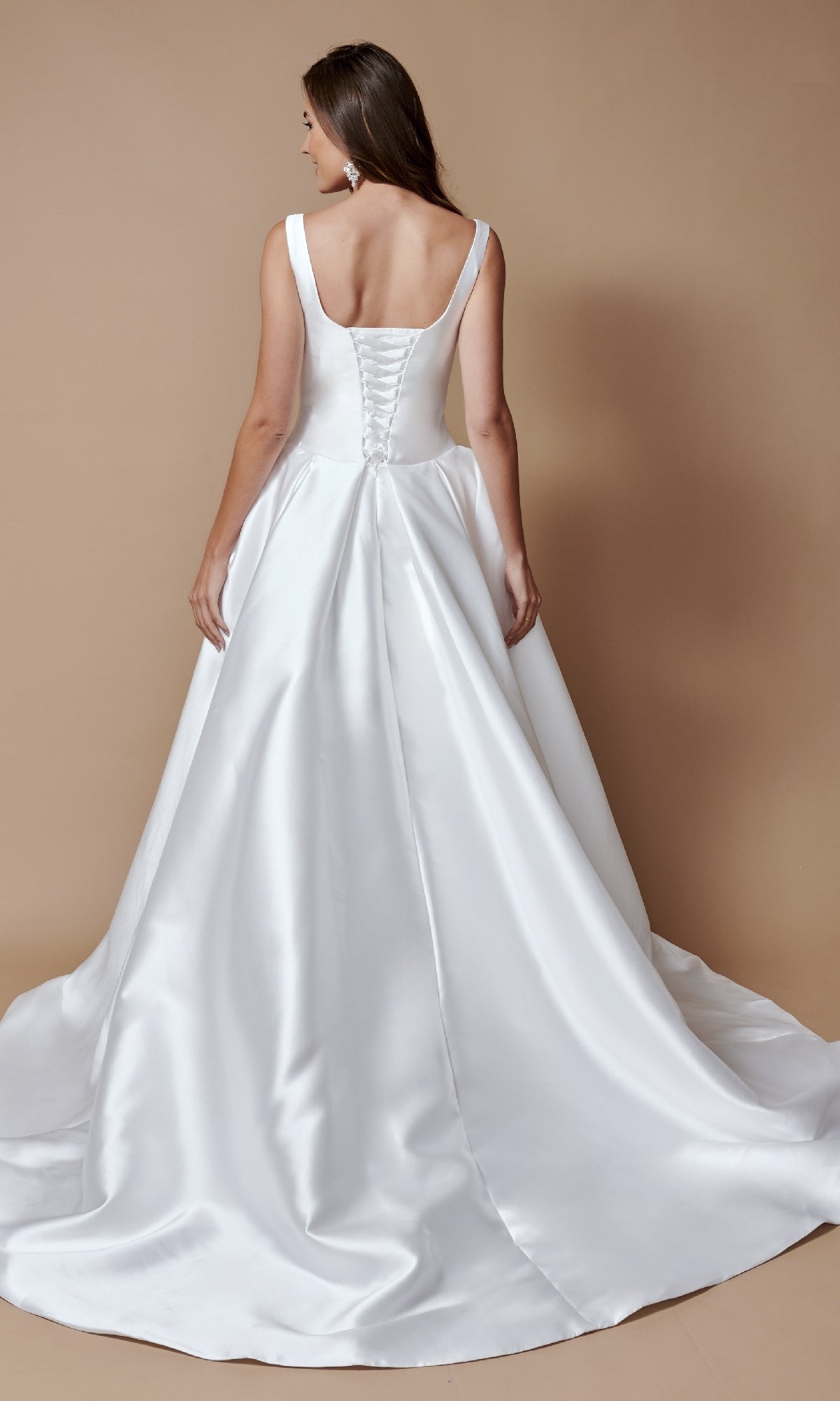 A woman models the Nina Canacci 3251 long formal dress—sleeveless satin gown with a lace-up back and flowing train—standing against a plain beige background.