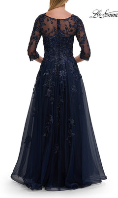 A woman is seen from the back wearing the La Femme 32538, an A-line navy blue tulle formal dress with sheer sleeves and delicate beaded lace appliques.