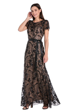 A woman models the R&M Richards 3253 Short Sleeve Black Formal Dress, featuring floral lace, a sheer overlay, and a black ribbon belt—an elegant special-occasion look—standing against a white background.