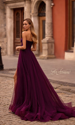 A woman wearing the La Femme 32579 Strapless Long Prom Ball Gown with a floral corset-style bodice and purple floor-length train stands on a cobblestone street, glancing over her shoulder.